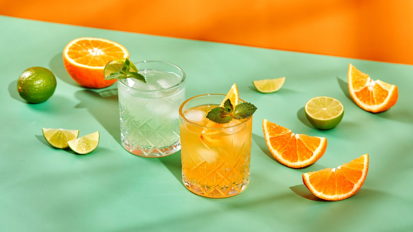 Multicolored cocktails in glasses of different shapes with orange and lime on colored backgrounds with hard shadows